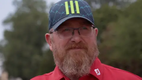 A close up of a man wearing a cap and glasses looking to the side of the camera. He is wearing a red shirt. 