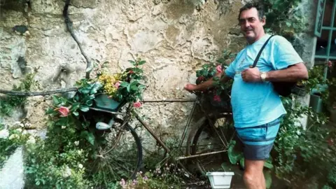 Family handout Derek Davies wearing a blue t-shirt and shorts standing next to a bike adorned with flowers in a house's front garden.