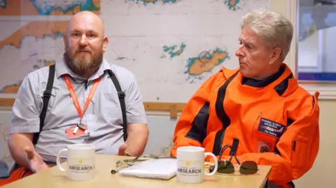 Two men sat at a table with a map of the Channel Islands behind them. One man is bald with a beard and is wearing a short-sleeved shirt and a lanyard. The other has grey hair and is wearing a bright-orange protective outfit for swimming / diving at sea.