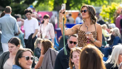 @SlowMoving Media A woman in a leapordskin dress and sunglasses takes a selfie in one of the open air areas at the Cheltenham Jazz Festival