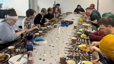 Thirteen people sitting at a long table eating Christmas dinner. There is a festive table cloth, brown paper cups, and plates full of food. There is a small Christmas tree behind one of the diners at the far end on the left.