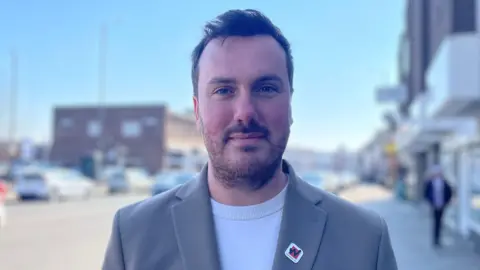 BBC/Gemma Dillon A man with short brown hair and a beard, wearing a grey suit jacket over a T-shirt.