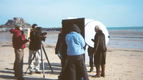 ArtHouse Jersey Film crew and cast stand on a beach while filming. A camera man is operating a camera which is pointing at cast members. It's a sunny day.