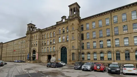 Google A large, multi-storey historical stone building with rows of tall windows, decorative arches, and small tower-like structures along the roofline. In front of the building, several cars are parked on a wet, paved area that appears to be a car park or loading space. The sky is overcast, and the overall scene has a quiet, subdued atmosphere with no visible people.