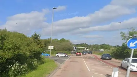 Google Cars queue at the junction of Whitley Wood Lane and the B3270. Large trees and grass are on either side the roads and a bridge can be seen in the distance