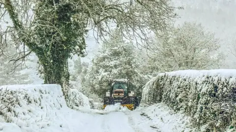 BBC Weather Watcher | Ruth Davies Tractor comes up a lane in Llanfair Dyffryn Clwyd in Denbighshire, surrounded by white snow. 