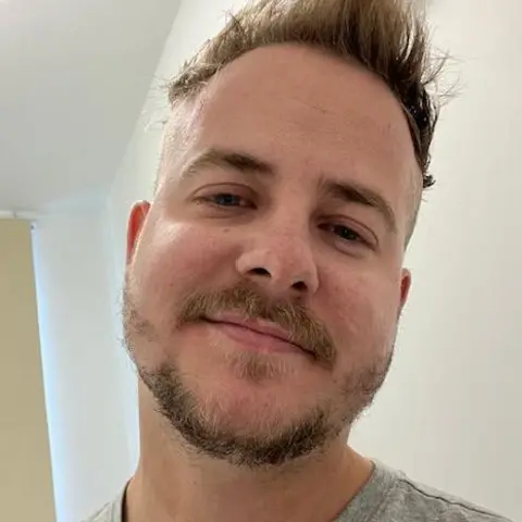 James Deegan James wearing a light grey T‑shirt standing indoors in a bright room. Part of a doorway or wall panel is visible behind him, with a window and a ceiling light in the background.