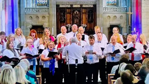 Hartlepool Youth Choir The Hartlepool ex-Youth Choir during one of their concerts. There are three rows of singers, all with lyric books in their hands and smartly dressed, singing in a church. A conductor has his back to the camera.