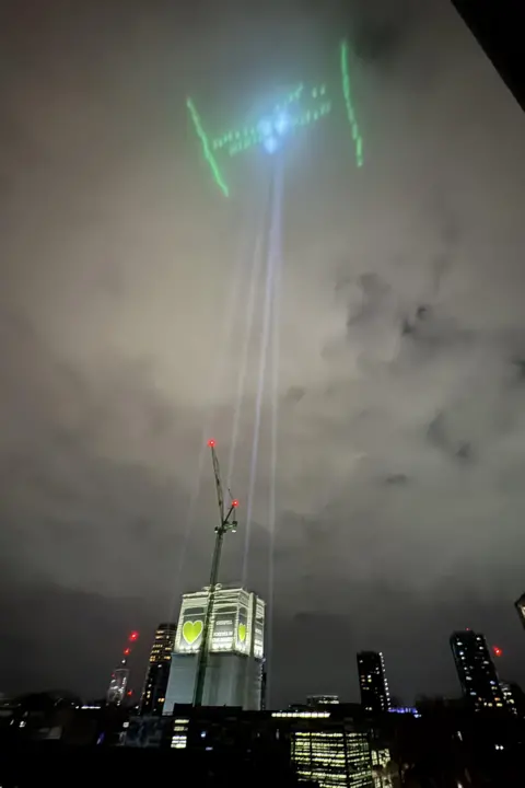 Reddit/@According_Appeal_654 Grenfell lit up in green with beams shining towards the sky. It is a cloudy night with a skyline of buildings in the background