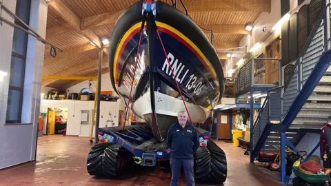 Dave is standing in front of the lifeboat inside Hoylake Lifeboat station. The lifeboat is towering over him and has RNLI 13-06 painted on it in white. Dave has his hands behind his back and he is smiling.