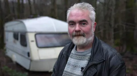 A man in his mid 50s with grey hair and a grey beard, wearing a leather jacket and patterned jumper. He is standing outside in front of a caravan. 