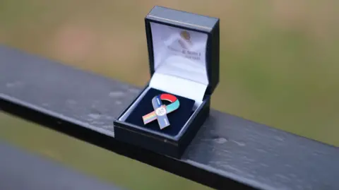 Ann Gannon / BBC The pin badge, or ribbon, on display in a small box, is ribbon shaped. It has a rainbow on one edge and is otherwise red, dark blue and light blue.