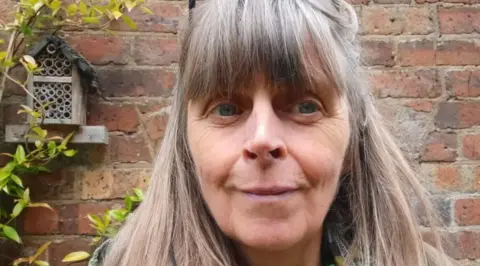Emma Purshouse A woman with long greying hair in front of a brick wall with a branch running up it