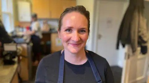 Ellie Rymer is a young woman with brown hair. She is wearing a chef's apron and dark T-shirt. She's smiling at the camera and is standing in a kitchen. 