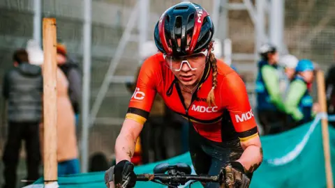 Pitchside Photo Lauren Fox, wearing a helmet, orange cycling top and black cycling shorts, looks determined as she guides her bike through a course. 