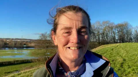 Amy Wheelton on her farm in Derbyshire with the River Trent in the background. Patches of green algae can be seen on the water with trees behind them. She has dark brown hair pulled back and is smiling at the camera, showing her top teeth. She has a cloth tied around her neck and wears a blue shirt with a green waistcoat over the top.