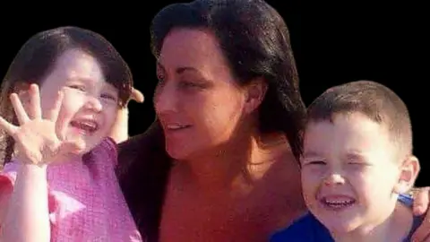 A woman with long black hair poses for a picture with her arms around a little girl in a pink dress and a boy in a blue t-shirt. 