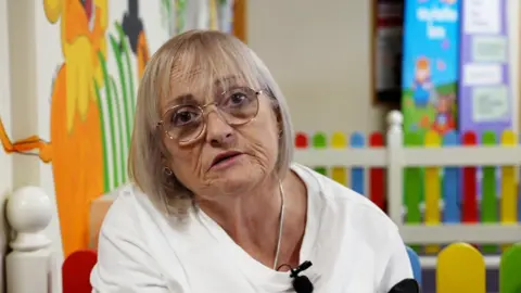 Karen McMillan is sat in a GP waiting room. She is wearing glasses and a white top.