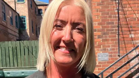 Phil Bodmer/BBC A blonde woman smiles at the camera. She is standing in a pub garden
