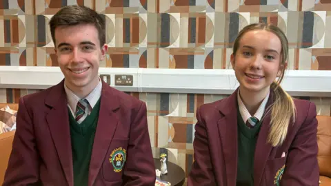 Two students, a boy and a girl, sitting side by side. The boy, with short brown hair, is sitting on the left side of the image and the girl, with blonde hair, tied up, is sitting on the right. They are wearing school uniforms, white shirts, dark green jumpers and burgundy blazers. 