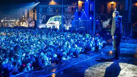 David Levene/Bradford City of Culture 2025 A man stands on on illuminated stage, with a crowd of thousands looking on. There's a blue hue over the whole scene and city centre buildings can be seen in the background.