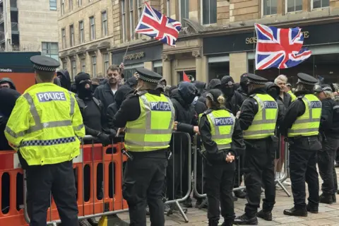 Police officers in high-visibility jackets stand in a line in front of metal crowd-control barriers, facing a group of people dressed mostly in black, some with face coverings, holding Union Jack flags outside city-centre buildings.