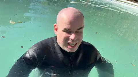 Simon Wilkes has a bald head and stubbly face and chin, he is wearing a wet suit and is in murky pool water, that has some leaves in it.