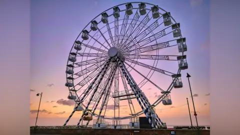 MrBlueSky/BBC Weather Watchers A large ferris wheel on the coast during sunrise. The sky is a mixture of orange, pink and purple colours. 