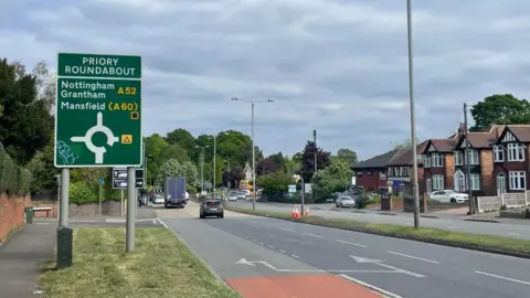 An image of the road leading up to the Priory Roundabout. A road sign in the foreground reads Priory Roundabout.
