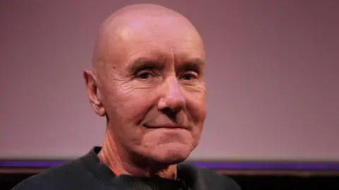 Irvine Welsh looks at the camera and smiles. He is bald and wears a dark top