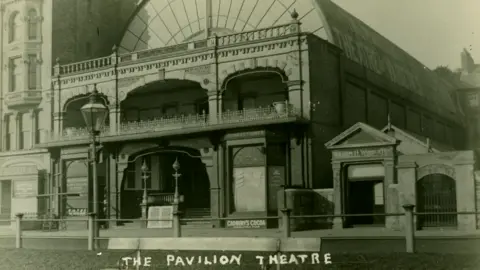 Manx National Heritage A black and white image of the Pavilion Theatre. The building had a semi-circular roof and a detailed iron frontage over two lower levels. You can see a small advert for Cadburys Cocoa by the entrance. At the bottom, the words The Pavilion Theatre have been written on the photograph in white.