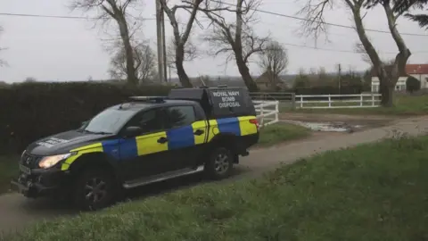 Rachel Holmes Royal Navy Bomb Disposal truck parked on a rural road with white fencing and a house seen in the distance.