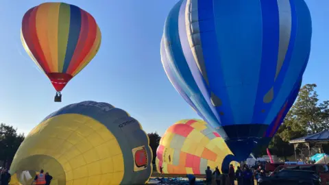 BBC Weather Watchers/Auper-skier A rainbow striped hot-air balloon is in the blue sky, while a large blue one is inflated but remains on the ground. Next to it is a yellow patchwork style balloon with blue, pink and orange squares. it is in the middle of being inflated and is lying on its side. Also being inflated on its side is a yellow balloon with a light blue strip, and a red shield on it.