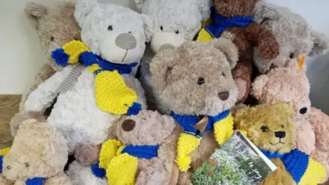 Ellie Somme A pile of bears are stacked together with yellow and blue scarves on indicating the colours of the Ukrainian flag.
