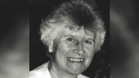 A black and white portrait photo of Beryl Purdy. She has short grey hair and is smiling at the camera. She is wearing a white top and pearl earrings.