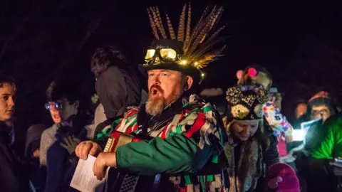 Tu Plus Tu Photography Simon processes from right to left with his mouth open as though proclaiming or singing. He wears an accordion on his chest and a feathered hat on his head. He has a green and red rag costume and holds papers that look like song lyrics. It is night time. People wearing winter coats and hats crowd around him. 