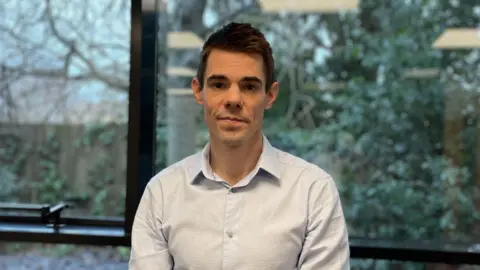 Dan Sexton is standing in an office room, wearing a light blue shirt and standing behind a window, in the background there are trees and bushes.