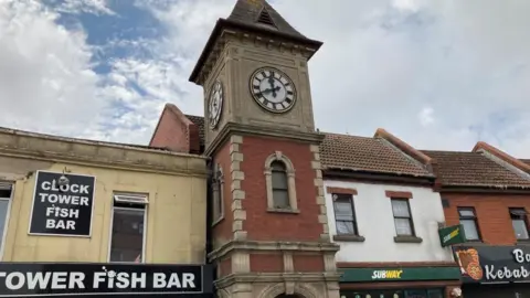 Kingswood clock tower is shown under cloudy skies. A sign on the front of the building to its left reads "Clock tower fish bar", while a Subway shop can be seen on the right.