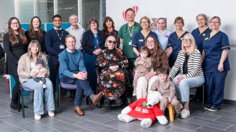 Children of the 90s A group of about 20 people posing for the camera with a seated row and others standing behind. Some are wearing hospital uniform and three mothers are with their young children