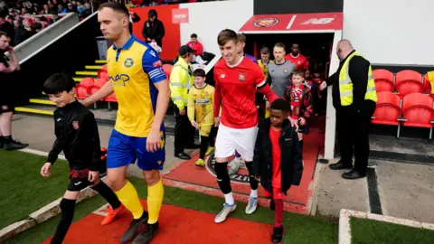 Players from Stockton Town FC and FC United of Manchester walk on to the pitch from the tunnel. Stockton Captain FC Adam Nicholson is leading his team, and is wearing the club's yellow and blue strip. He has arm on the shoulder of a ball boy walking besides him.