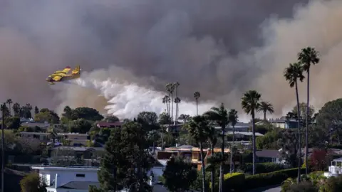 Getty Images A small plane drops water over buildings in a densely populated area, while smoke fills the area behind them