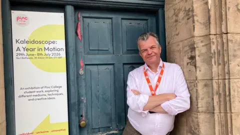 BBC A man in an open necked shirt leans against a stone pillar, in front of a wooden door with peeling paint. The man has his arms crossed and is smiling