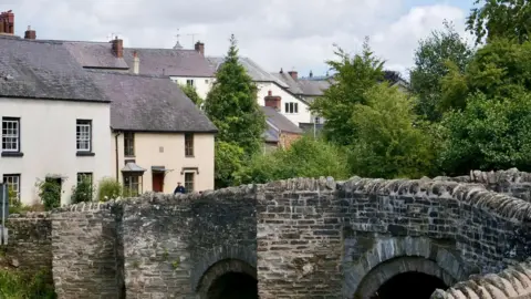 John Bray/BBC A row of white-walled houses with grey roofs with a stone bridge in the foreground