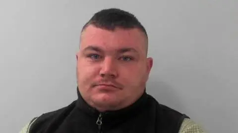 A mugshot of a young man with short hair and wearing a zip up black vest. 