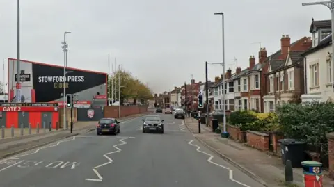 Google Kingsholm Road in Gloucester - a residential street with Edwardian terraced houses on the right and Kingsholm rugby stadium - which has black, grey and red cladding - on the left. There are two cars driving over a pedestrian crossing and the sky overhead is cloudy.
