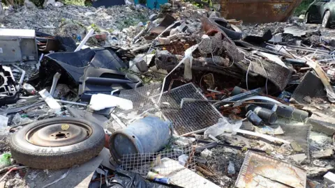 Defra A large pile of mixed waste and scrap materials is scattered across an outdoor industrial site. Visible items include a discarded vehicle tyre, metal mesh panels, twisted wire, broken furniture parts, metal cylinders, plastic containers, pipes, and assorted construction debris.