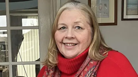 Supplied Joanne Thorns is wearing a red jumper with a high neck and a scarf on top. She has shoulder length light brown hair and is smiling inside a house. 