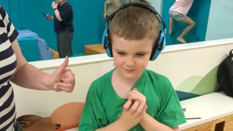 A child wearing blue Active Noise Cancelling headphones and wearing a green t-shirt.