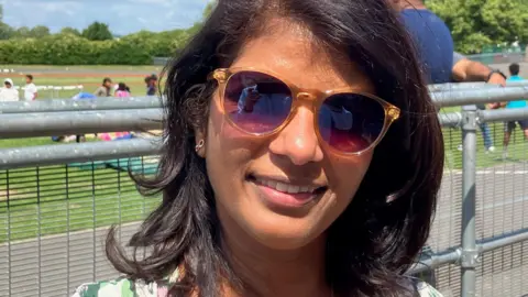 Ms Furtado is has wavy dark hair and is wearing sunglasses and a green patterned top. She's smiling at the camera and there's grass and a metal railing behind her. It's a sunny day and the sky is blue.
