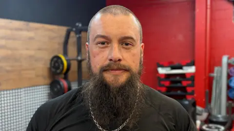 A man with a beard, shaved head, black t-shirt and a silver chain on.